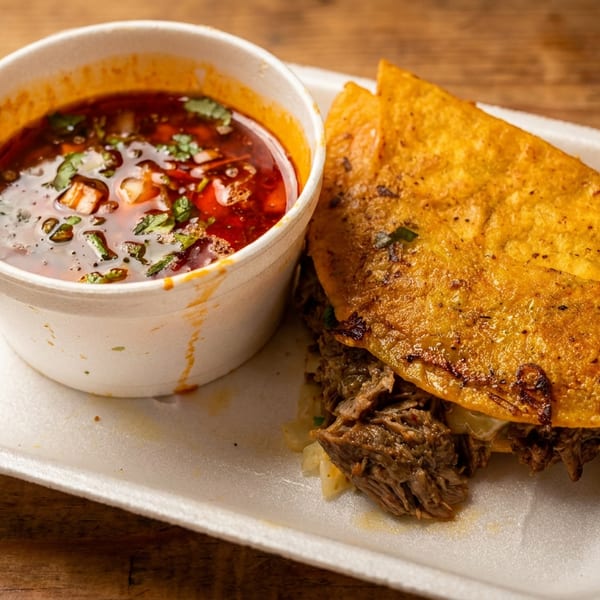 A birria taco with consommé in a white styrofoam container on a wooden table.